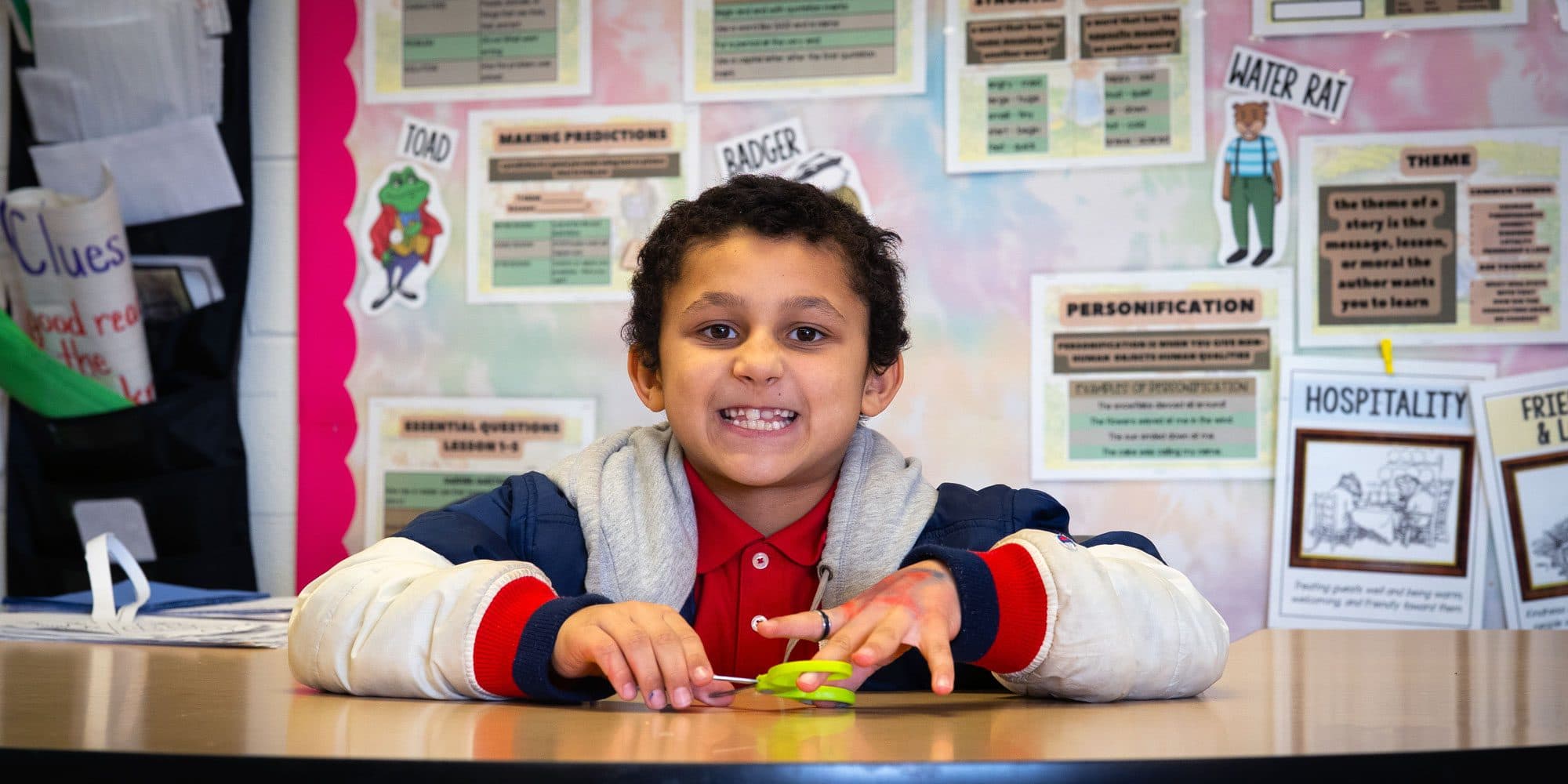 Smiling student seated at a desk in front of a reading-skills bulletin board.