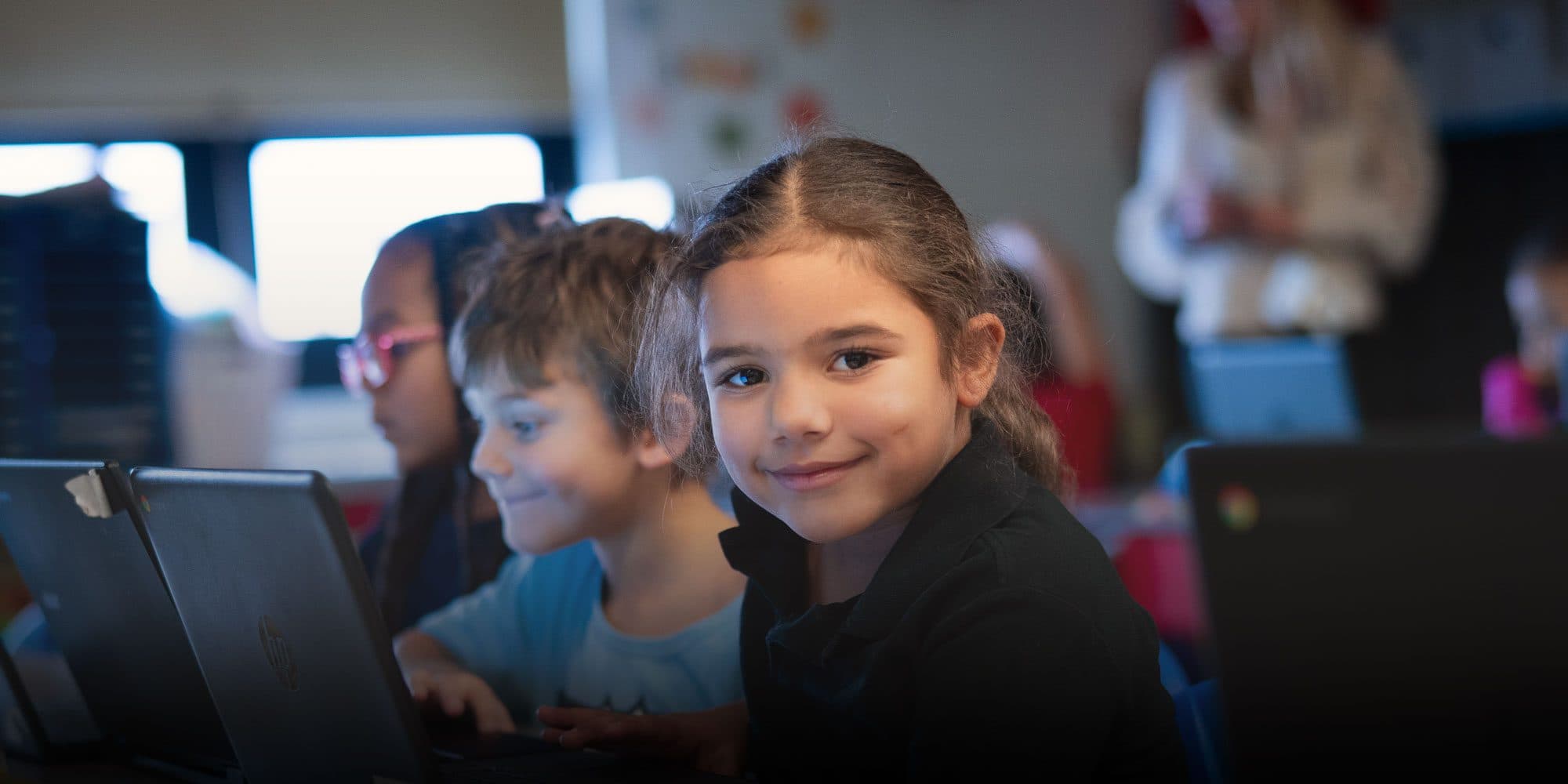 Smiling student at a laptop beside classmates in a classroom.
