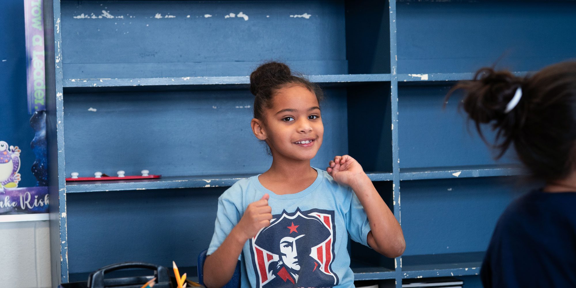Smiling student in a Patriots T-shirt in front of blue classroom shelves.