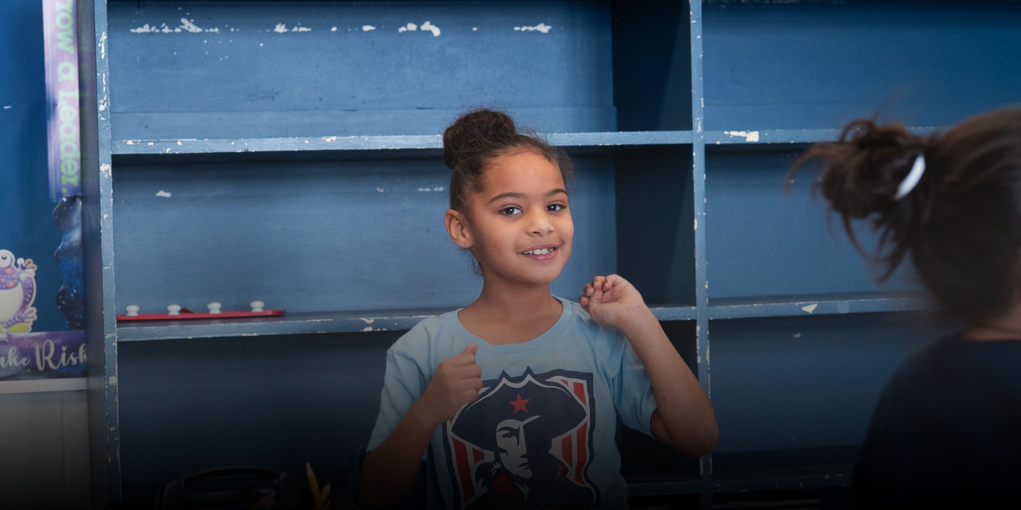 Smiling student in a Patriots T-shirt in front of blue classroom shelves.
